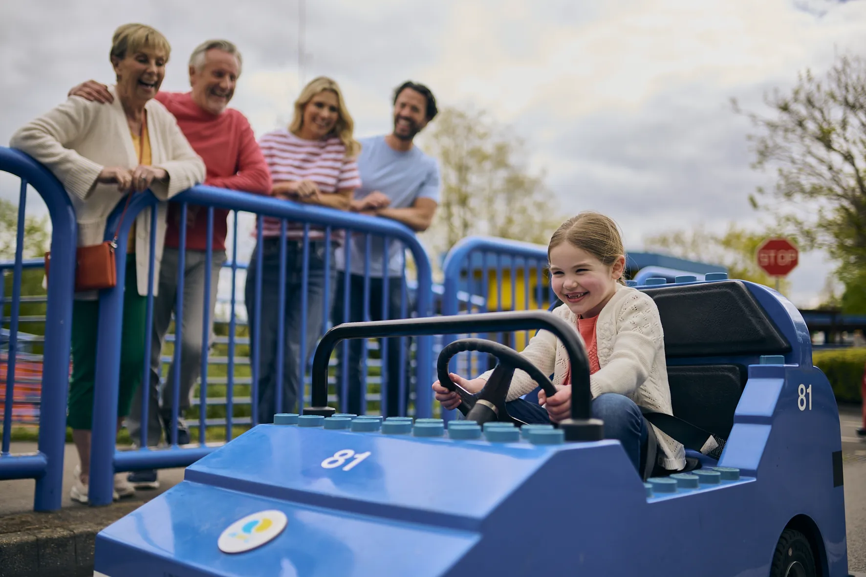 Young girl driving a LEGO City Car with her family smiling over at her in the background behind blue barriers 